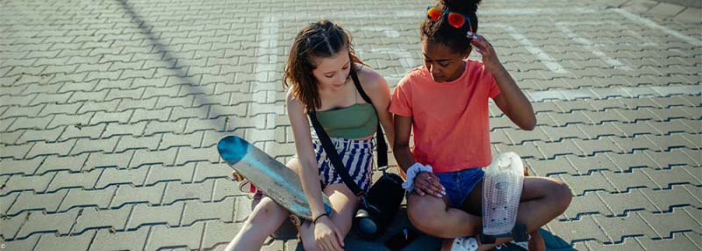 Foto: Zwei junge Mädchen mit Skateboards sitzen auf einem asphaltierten Platz und reden.