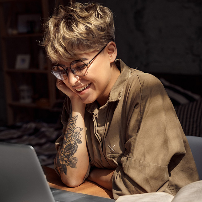 Foto: weiße, tätowierte, junge Frau mit kurzen Haaren und Brille sitzt vor Laptop und lächelt